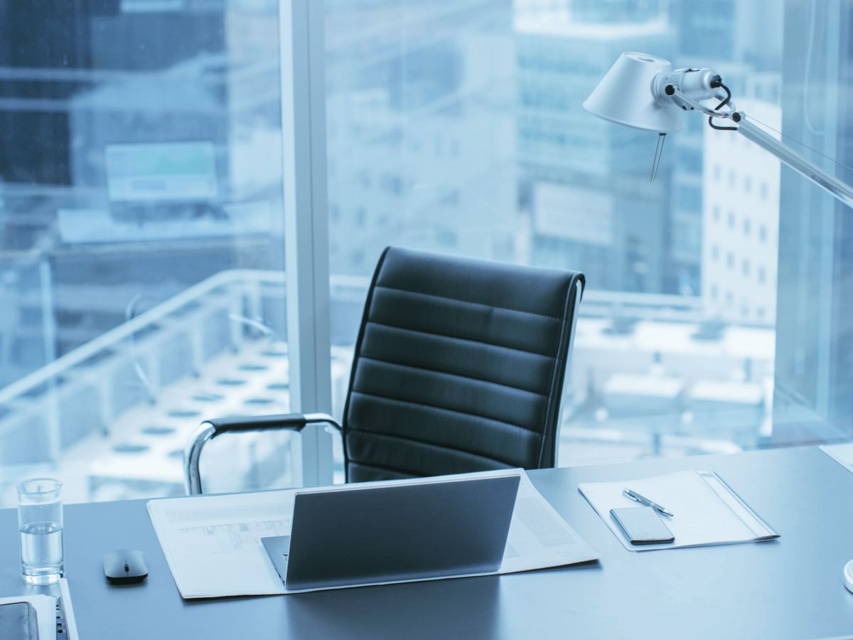 An empty black executive chair at a desk with a laptop and documents, beside a high-rise office window overlooking a city skyline